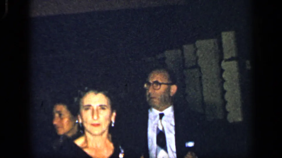 A woman in formal attire faces the camera while a man with glasses and a tie stands behind her in low-light 1948 New York home-movie footage.