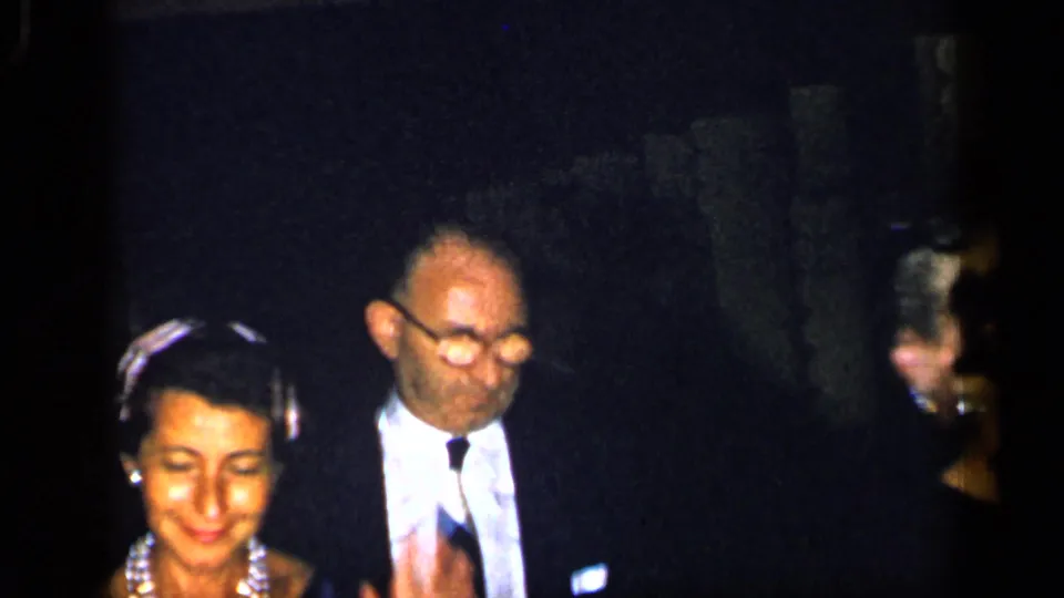 A woman wearing a decorative headpiece and earrings walks beside a man in glasses and a dark suit, with other formal guests partly visible in the 1948 New York scene.
