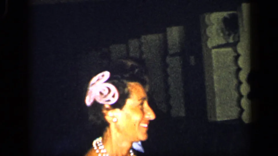 In the 1948 New York home-movie footage, a smiling woman with pearls and a pink hair accessory appears near a dark stage setting.