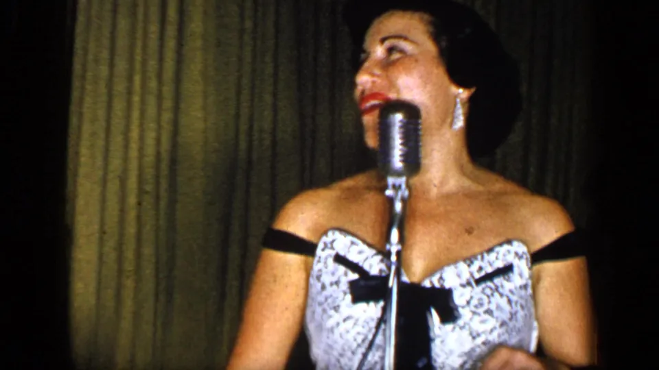 A woman in a patterned dress with a black bow sings into a chrome microphone on a dimly lit New York stage in 1948 footage.
