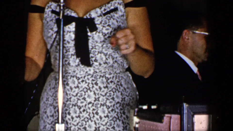 A woman performing at a microphone is framed from the waist up, with a suited man partly visible behind her in the 1948 New York footage.