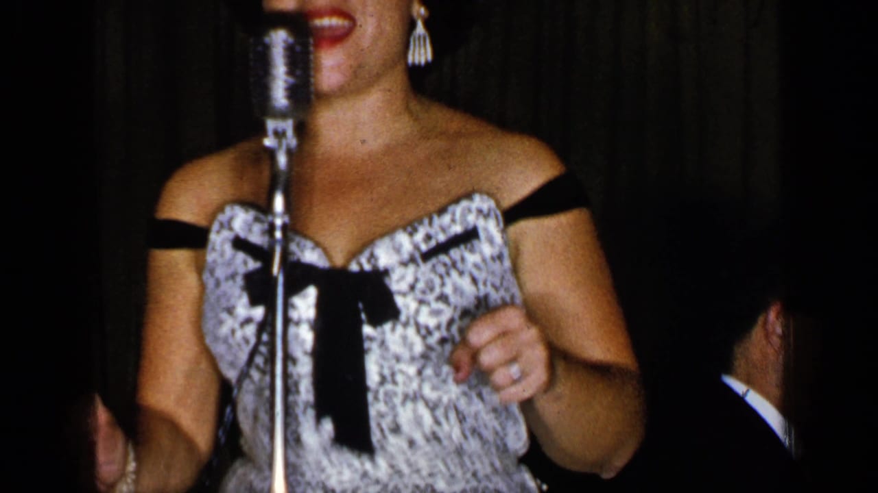 Woman in patterned off-the-shoulder dress singing at a standing microphone on a dim stage.