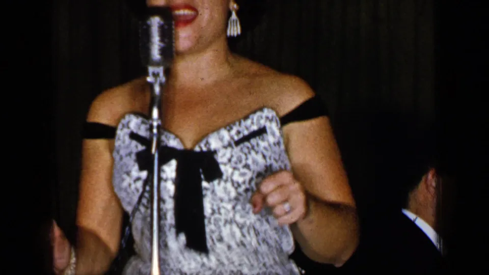 In 1948 New York footage, a woman in a patterned off-the-shoulder dress stands close to a vintage microphone during a stage performance.