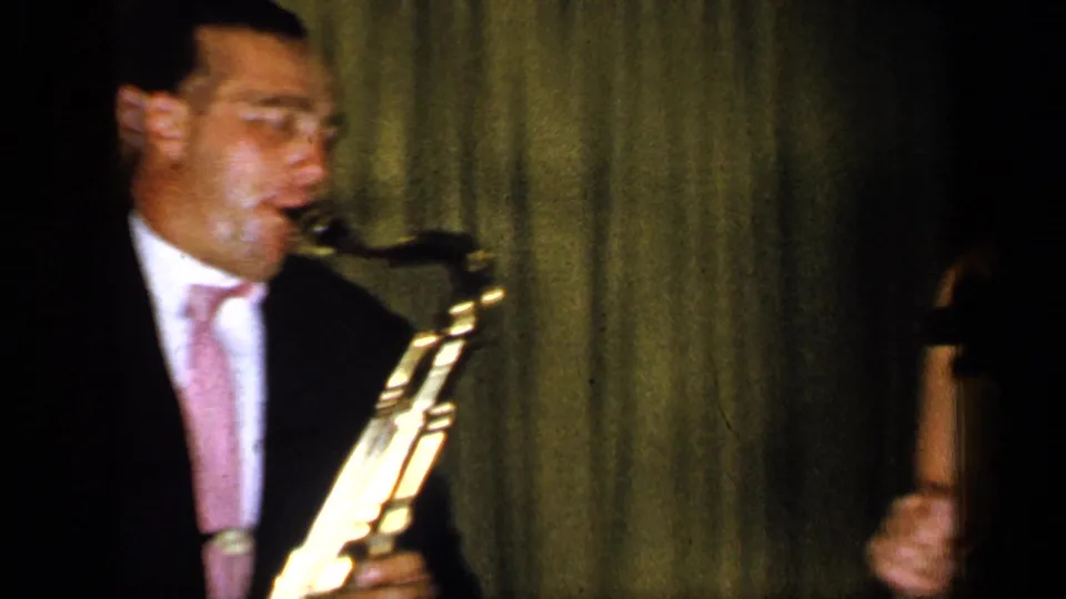 A male saxophonist in a dark suit and pink tie plays on a dim indoor stage, with curtain fabric behind him.