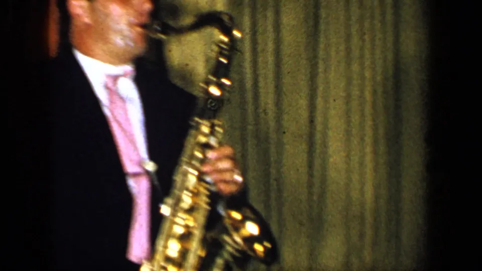 The saxophonist remains framed close-up as the brass instrument catches light against the dark indoor performance backdrop.
