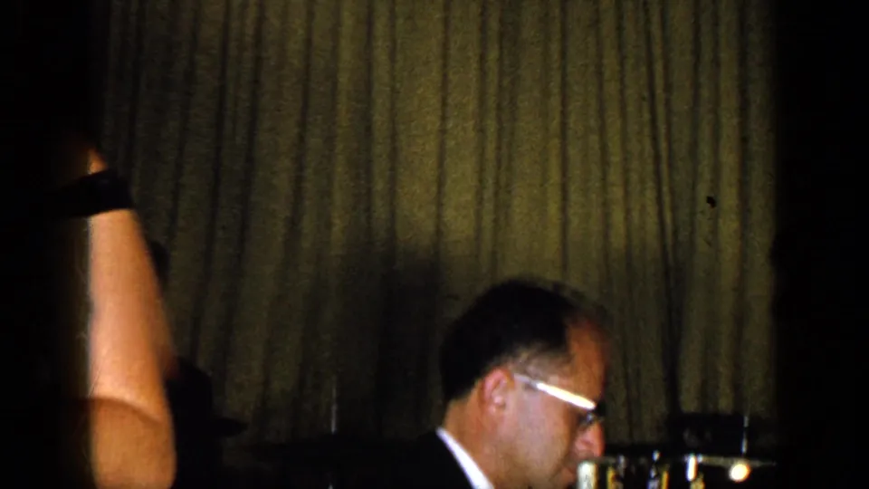 A cropped stage view shows part of the singer at left and a seated man wearing glasses near instruments against the curtain backdrop.