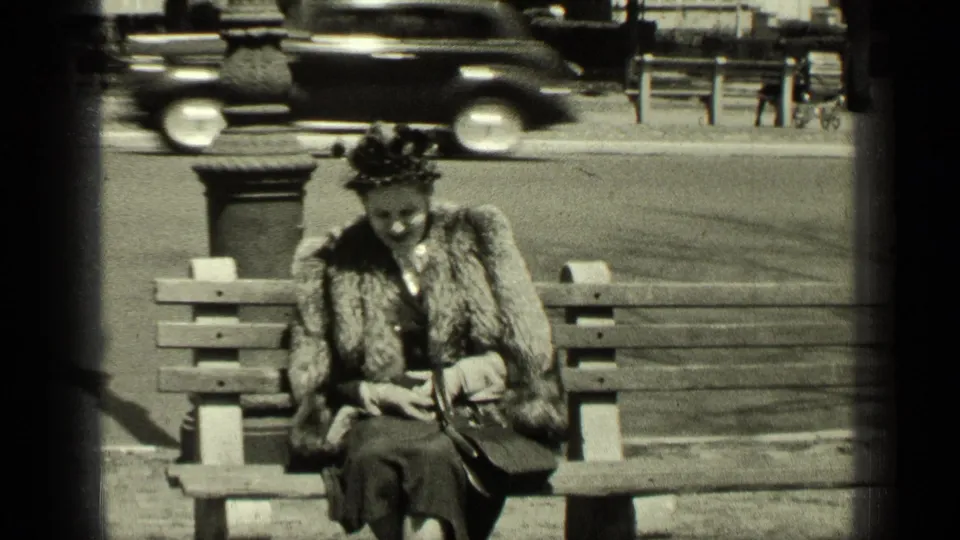 In 1949 New York, a woman in a fur stole and vintage hat sits on a park bench while a car passes on the street behind her.