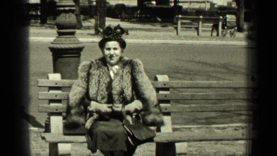 The woman sits upright on a New York park bench, facing the camera with a fur stole, hat, gloves, and handbag visible.