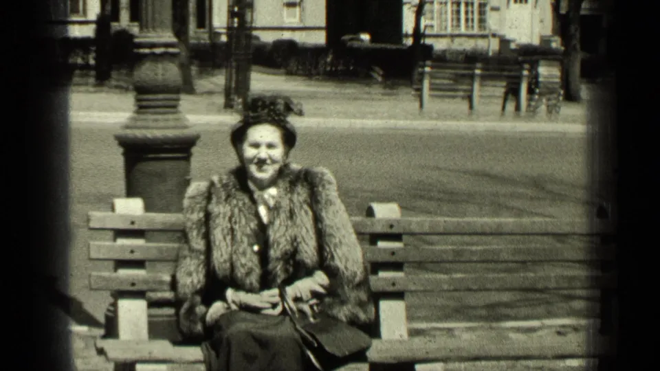 A woman in late-1940s dress smiles from a park bench in New York, with her handbag at her side and street details behind her.