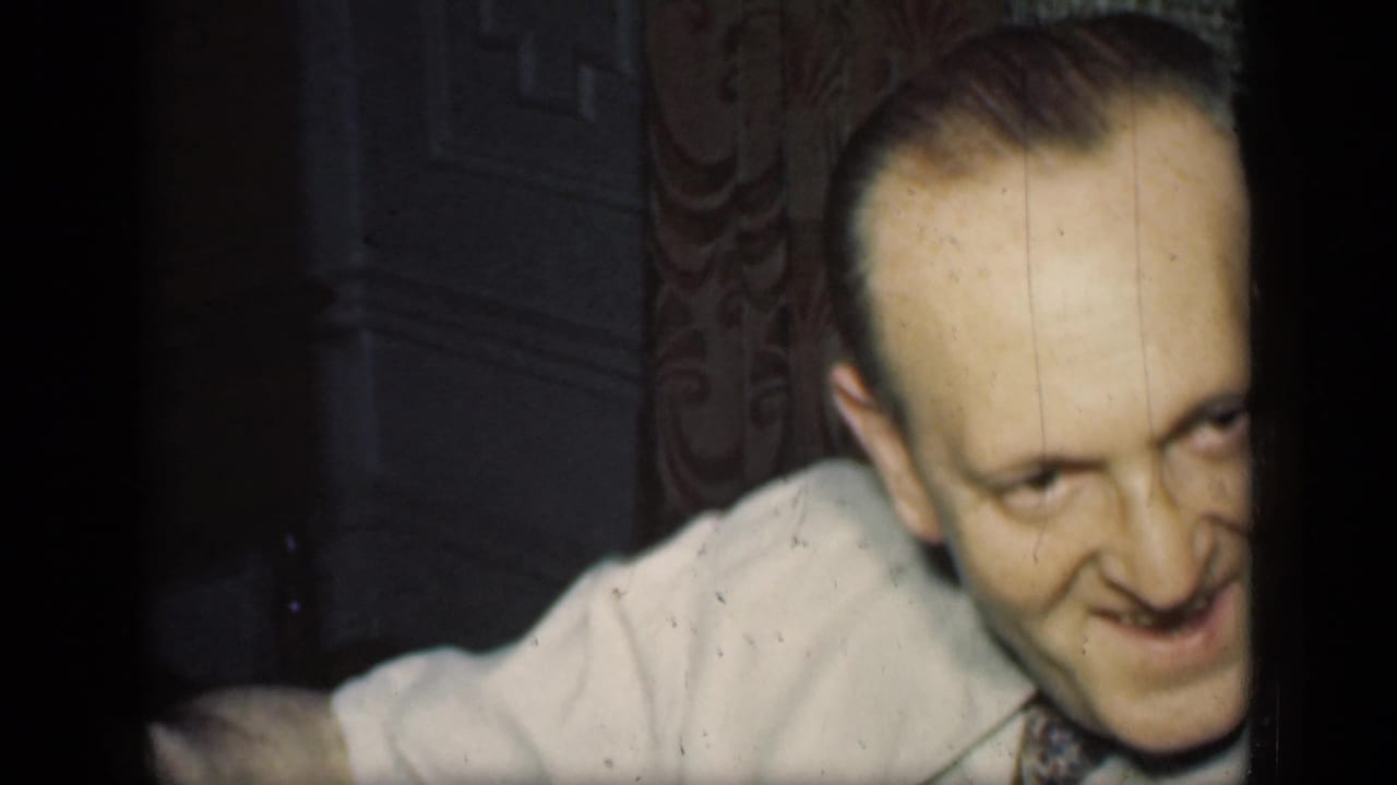 Smiling man in a light shirt and tie leans forward in a dim room with patterned wallpaper behind him.