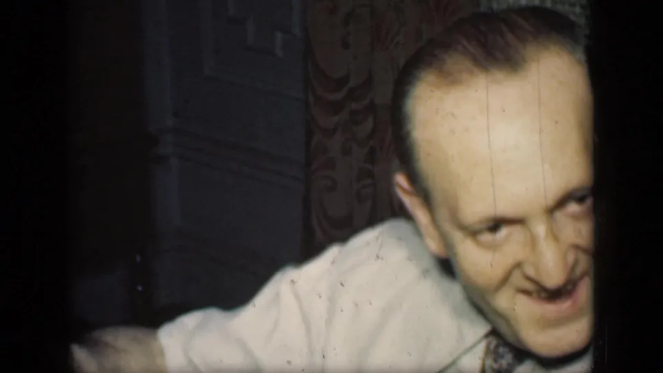 A smiling man in a light dress shirt and patterned tie leans toward the camera in a dim 1949 New York home interior.