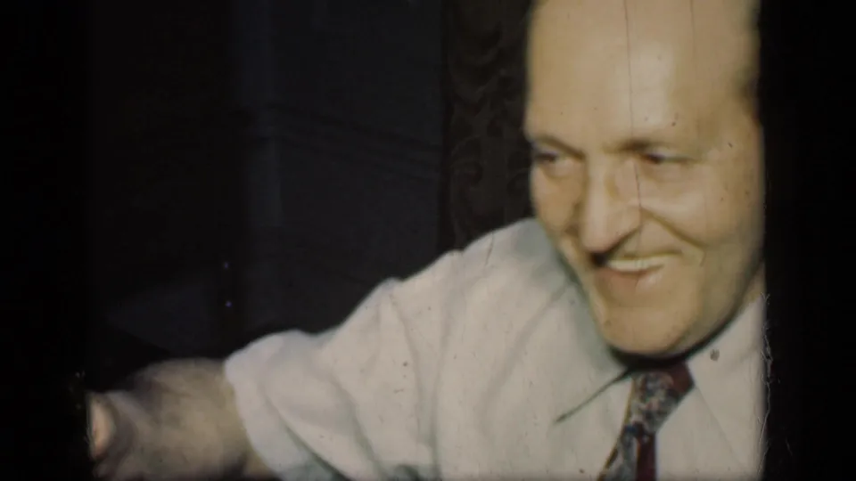 The man smiles broadly in a close 8mm frame, with a light shirt, patterned tie, wallpaper, and a dim domestic room visible.