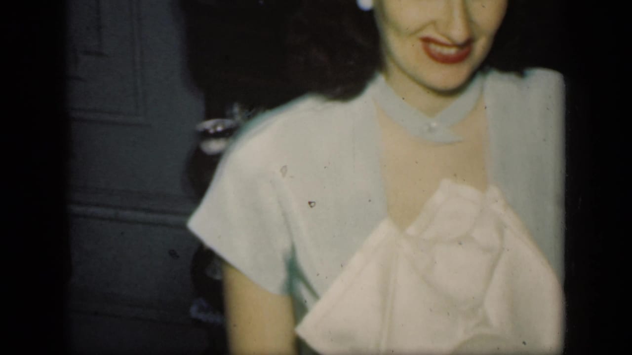 Adult woman in light clothing smiles in a dim New York home-movie interior, with her face partly cropped by the frame.