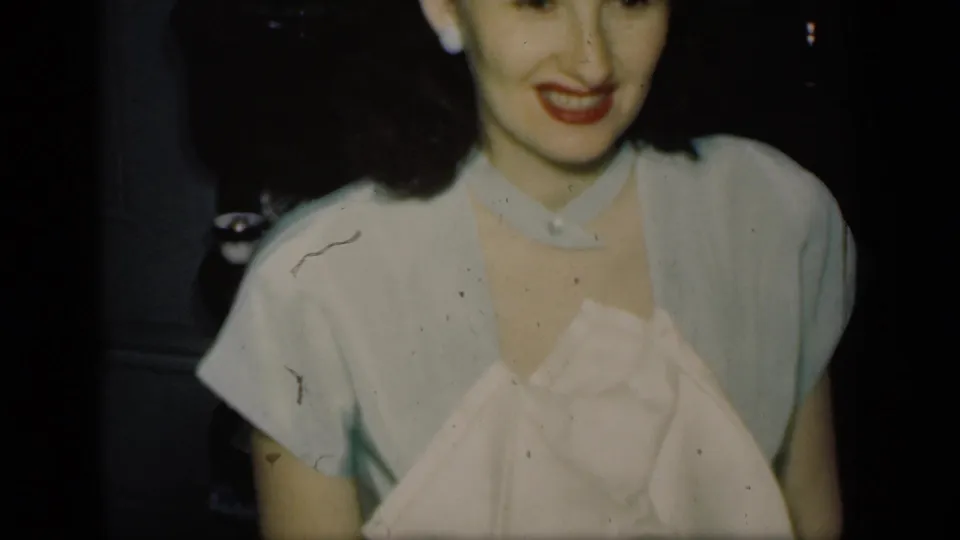 In this 1949 New York home-movie still, a woman smiles toward the camera while seated or standing in a dim private room.