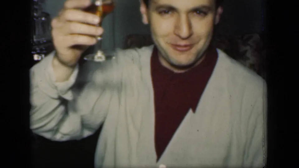The man faces the camera with a raised wine glass, wearing a light cardigan in a dim 1949 New York dinner scene.