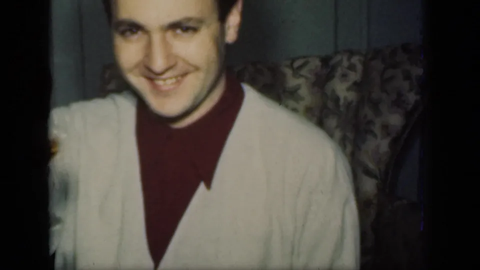 A smiling man in a light cardigan sits near upholstered seating in the dim 1949 New York dinner setting.