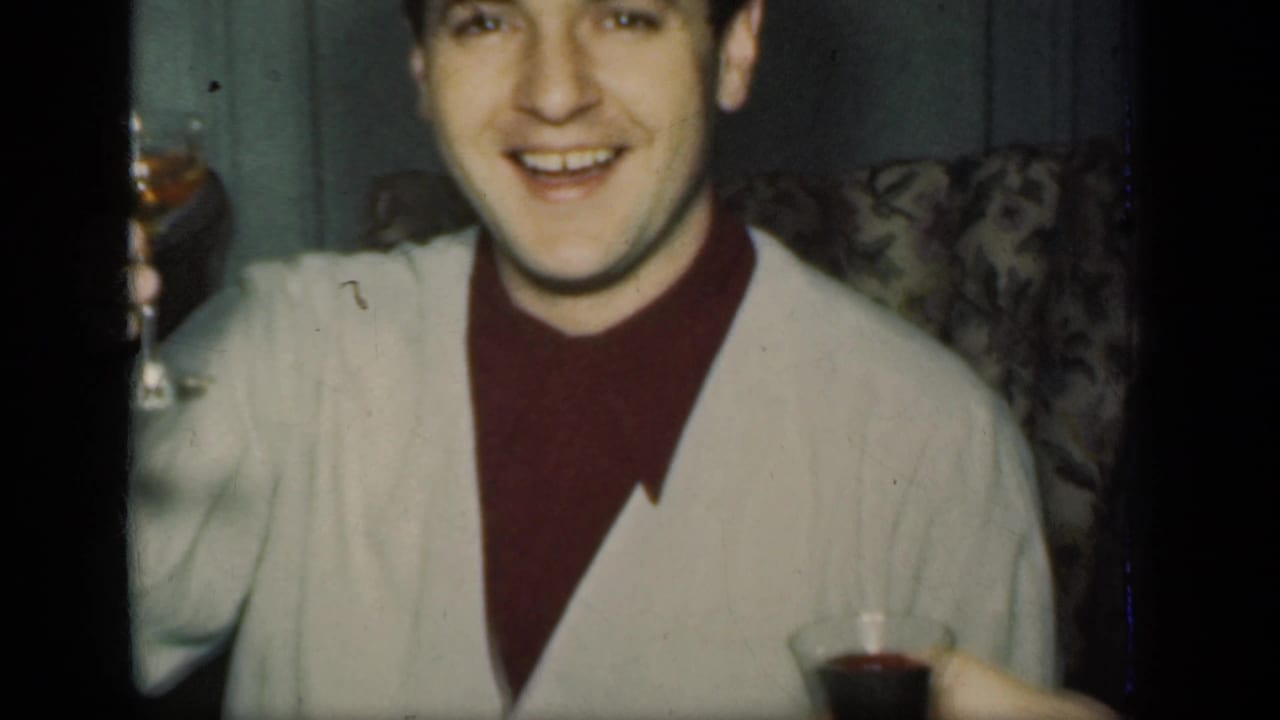 Smiling young man in a white cardigan holds a glass in a dim indoor room.