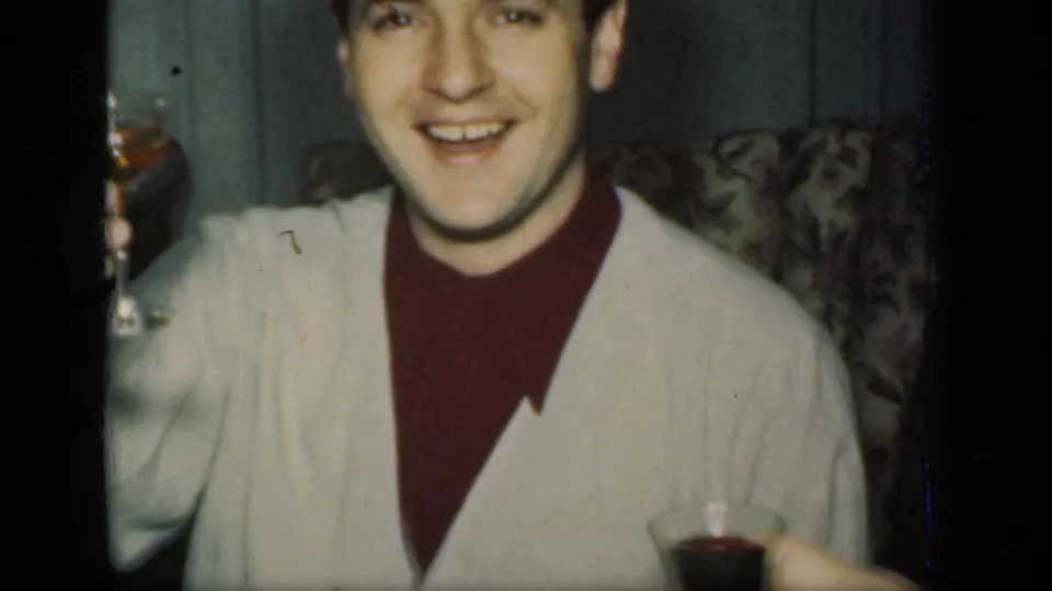 In a 1949 New York home-movie frame, a young man in a white cardigan and maroon turtleneck smiles while holding a drink indoors.