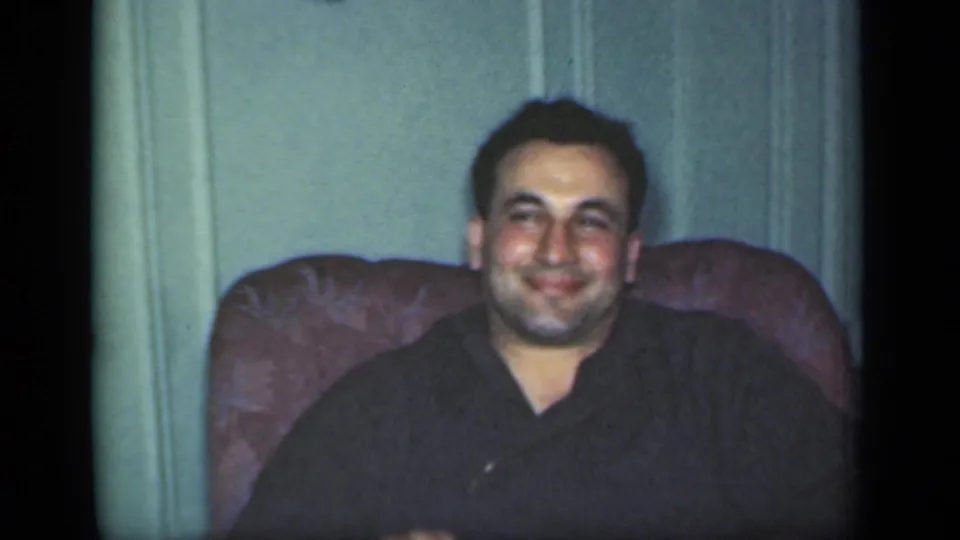 A man in a dark shirt smiles from an upholstered chair in a 1949 New York home-movie interior.