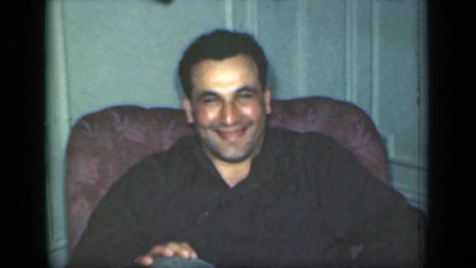 A seated man smiles with clasped hands in a private New York interior, shown in soft 8mm color from the 1949 clip.