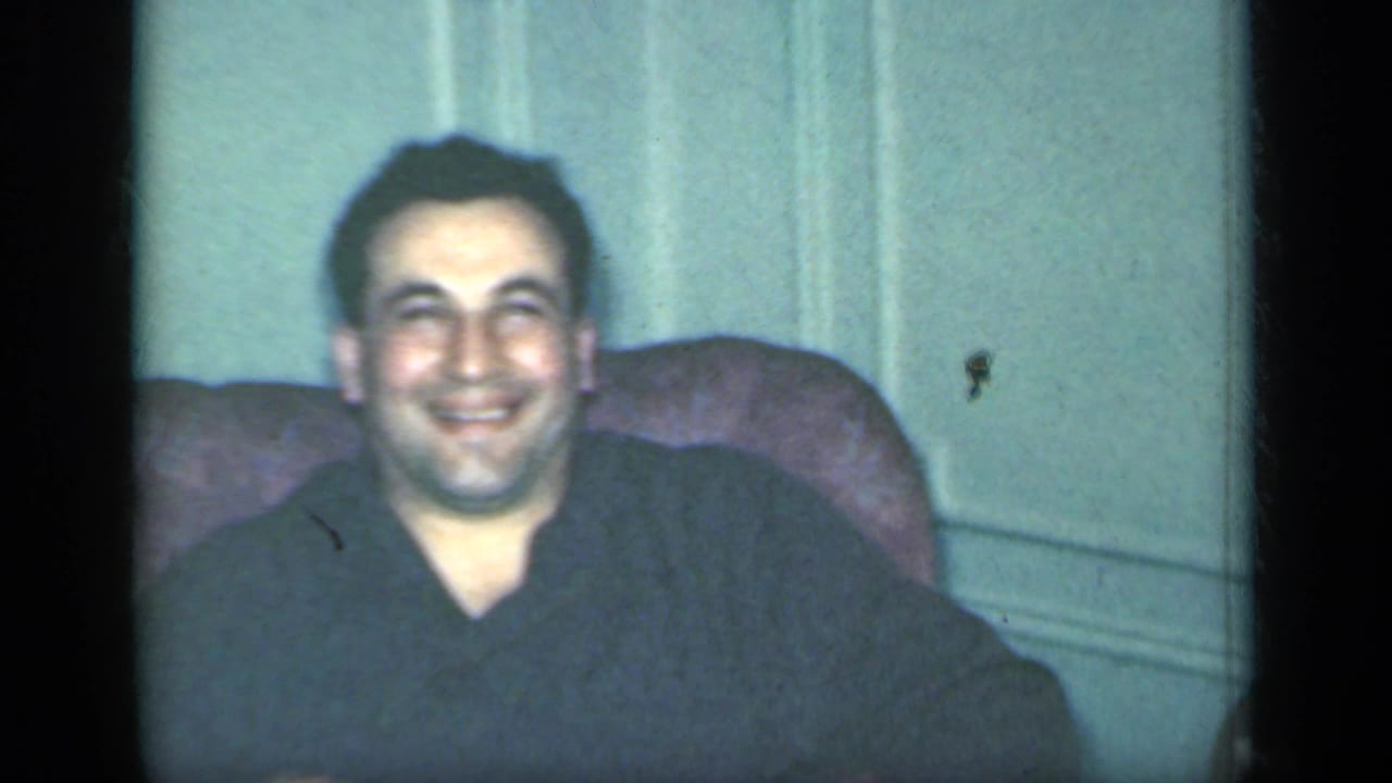 Smiling man seated in an armchair beside a pale interior wall.