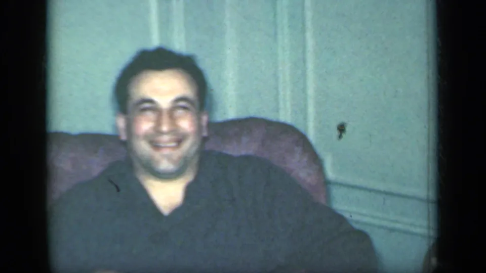 In 1949 New York home-movie footage, a man in a dark top smiles from a patterned armchair in a private interior.