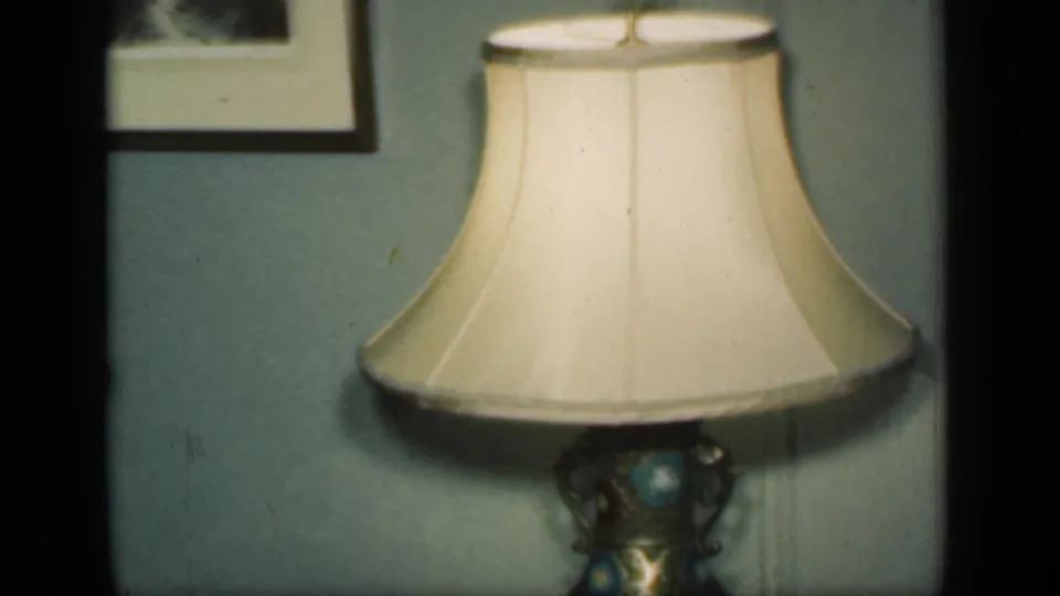 A cream lampshade fills the lower frame while framed wall art and pale interior wall remain visible in the home-movie scene.