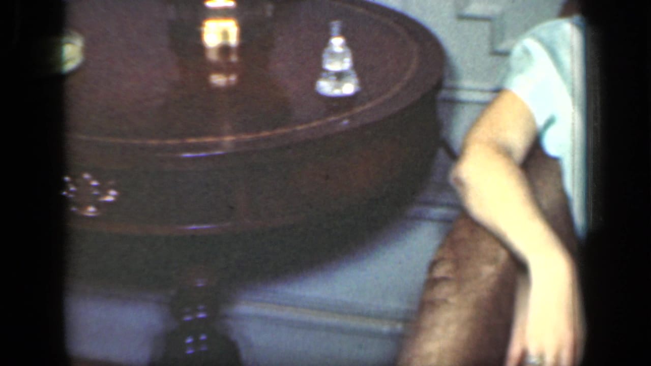 1949 New York interior frame with a round wooden table, small bell-like object, and the arm and skirt of a seated woman.