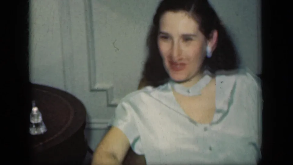 1949 New York home-movie frame showing an adult woman with dark styled hair, pale blouse, and earrings seated in a living room.