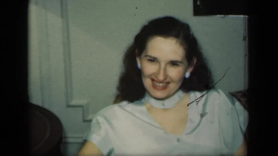 1949 New York frame of a smiling adult woman in a pale blouse and earrings, seated in a private living-room interior.