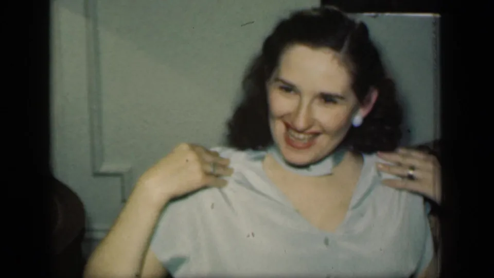 In a 1949 New York home-movie frame, a woman smiles toward the camera while seated on a brown sofa in a light blue blouse and neck scarf.