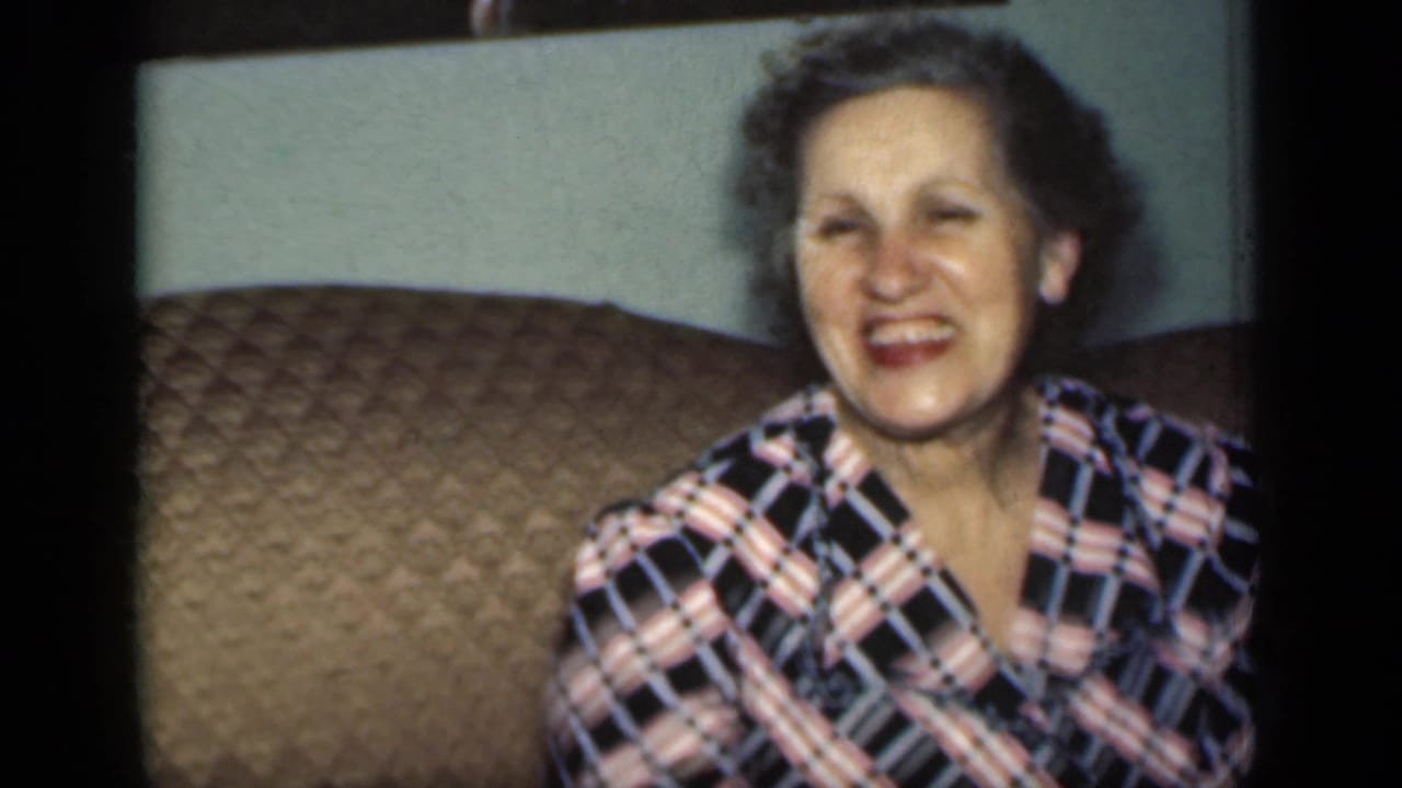 Smiling older woman seated on a brown quilted couch against a pale wall.
