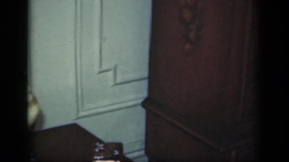 1949 New York home-movie frame showing a dark wooden wardrobe near pale wall paneling, with soft 8mm grain and partial shadowing.