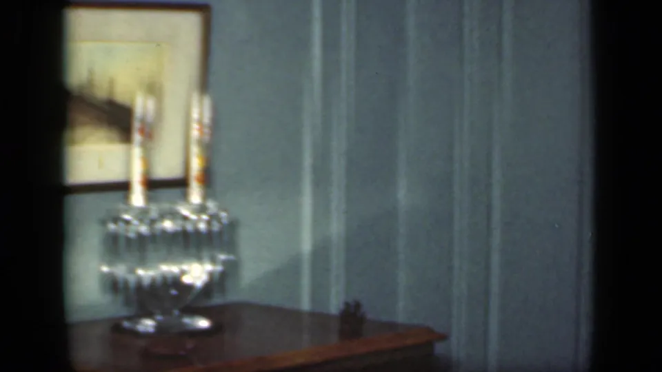 The frame shows a glass candle holder with candles, a framed mirror, and paneled wall details in the 1949 New York home interior.