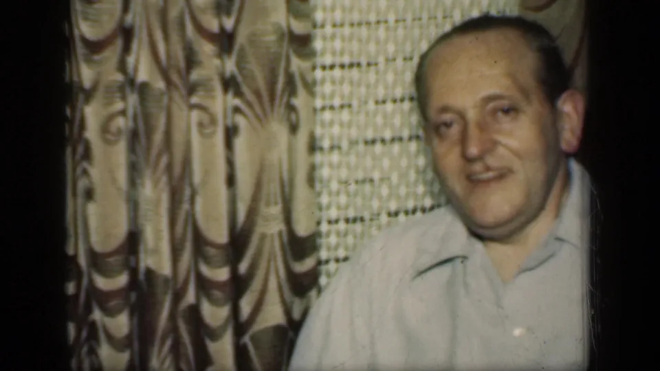 1949 New York home-movie frame of an adult man in a light collared shirt near patterned curtains and a lace window covering.