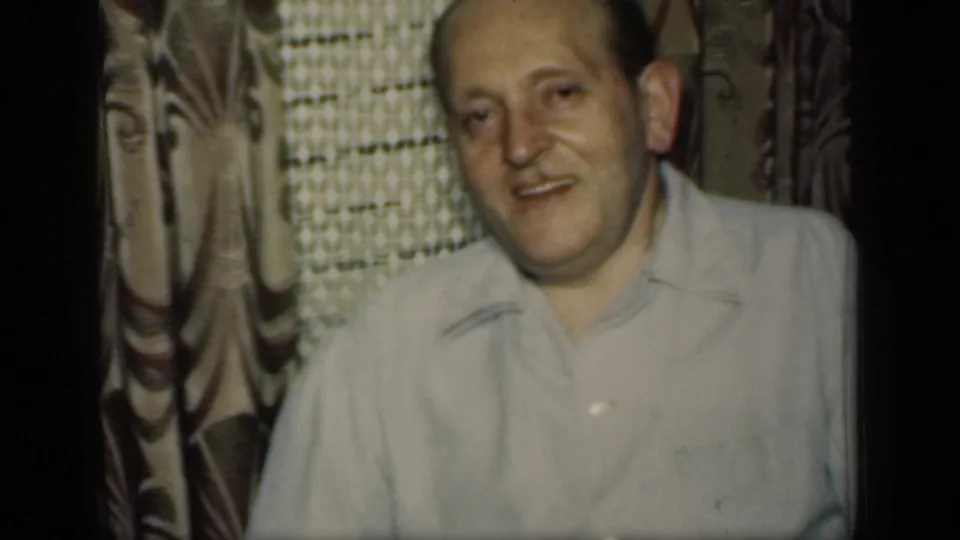 1949 New York home-movie frame showing a smiling adult man in a light collared shirt in front of patterned curtains.