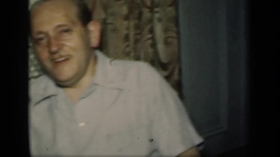 In a 1949 New York home-movie frame, an adult man smiles toward the camera near patterned decor and pale interior trim.