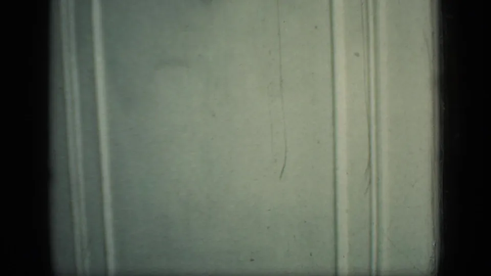 A close 8mm frame shows pale interior wall paneling in the New York home, with film grain and slight vertical marks visible.