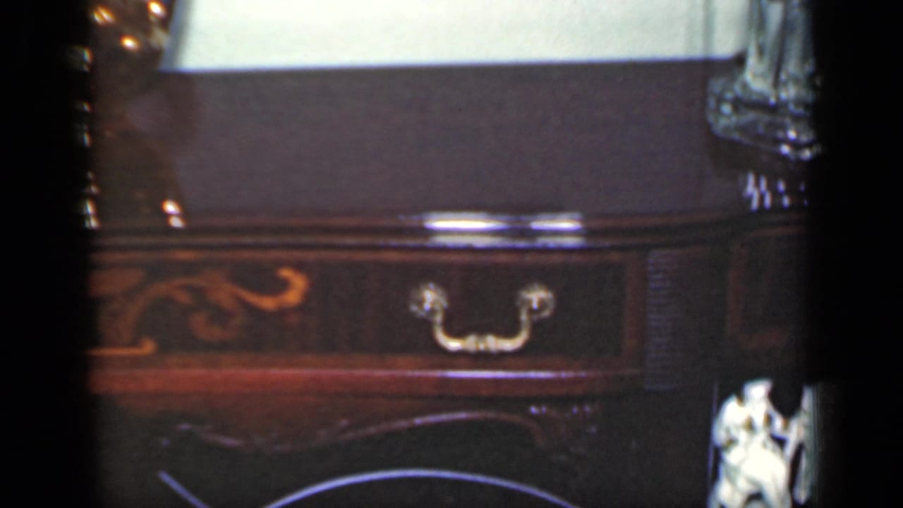 1949 New York 8mm frame showing a dark wood console table with brass hardware, inlaid detail, and a pale paneled wall.