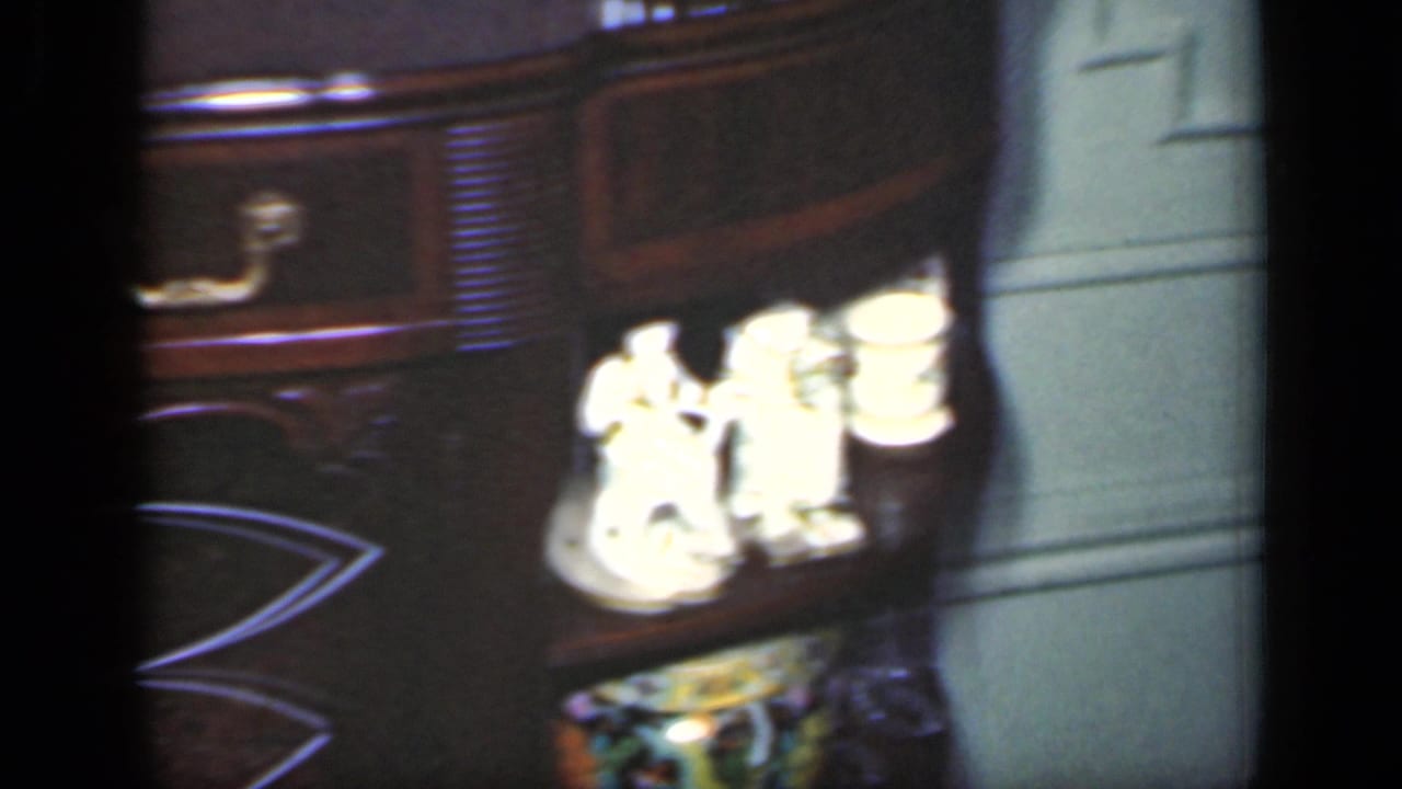 1949 New York home-movie frame of a console table corner, white figurines, brass hardware, and pale paneled wall.