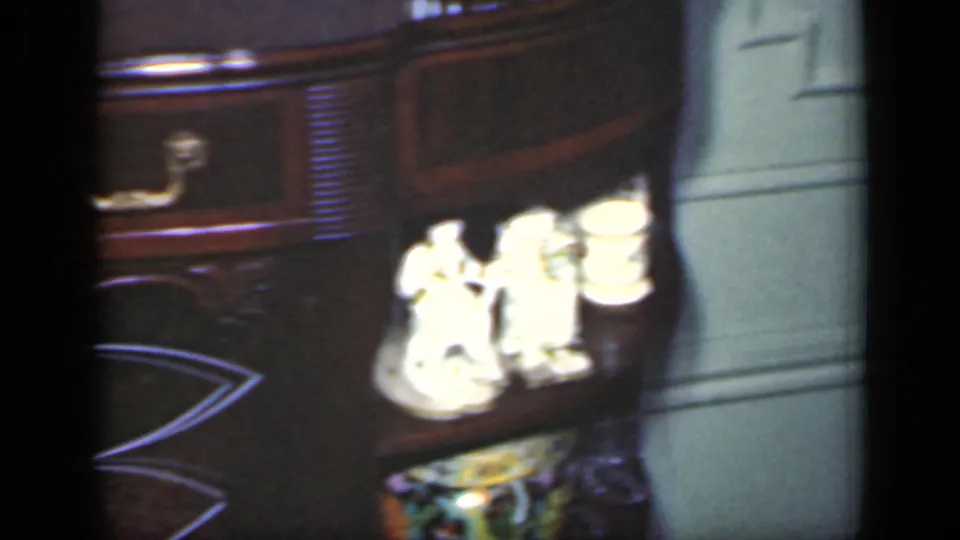 1949 New York home-movie frame of a console table corner, white figurines, brass hardware, and pale paneled wall.