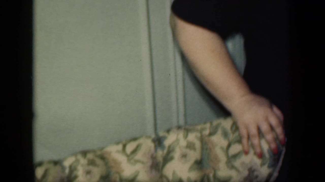 Partial view of a woman resting a hand on an upholstered chair in a dim interior.