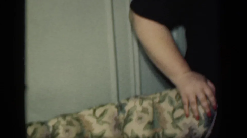 In a 1949 New York home-movie frame, a partially seen woman in dark clothing rests her hand on patterned upholstery near a pale wall.