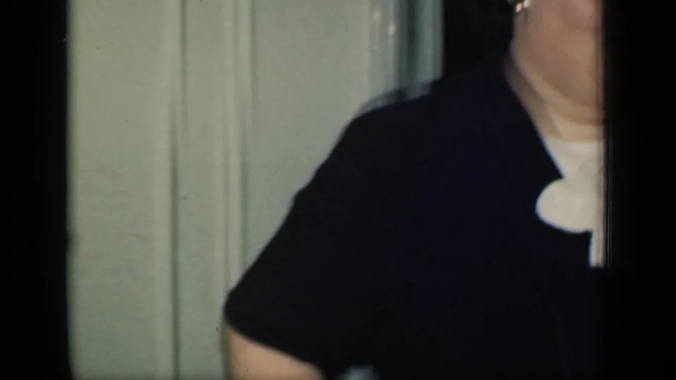 A 1949 New York home-movie frame shows part of a woman in a dark jacket with a white ruffled collar beside a pale wall and dark doorway.