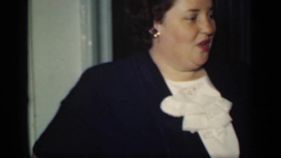 In this 1949 New York interior frame, a woman wearing a dark jacket, white ruffled collar, and gold earrings looks off to the side.