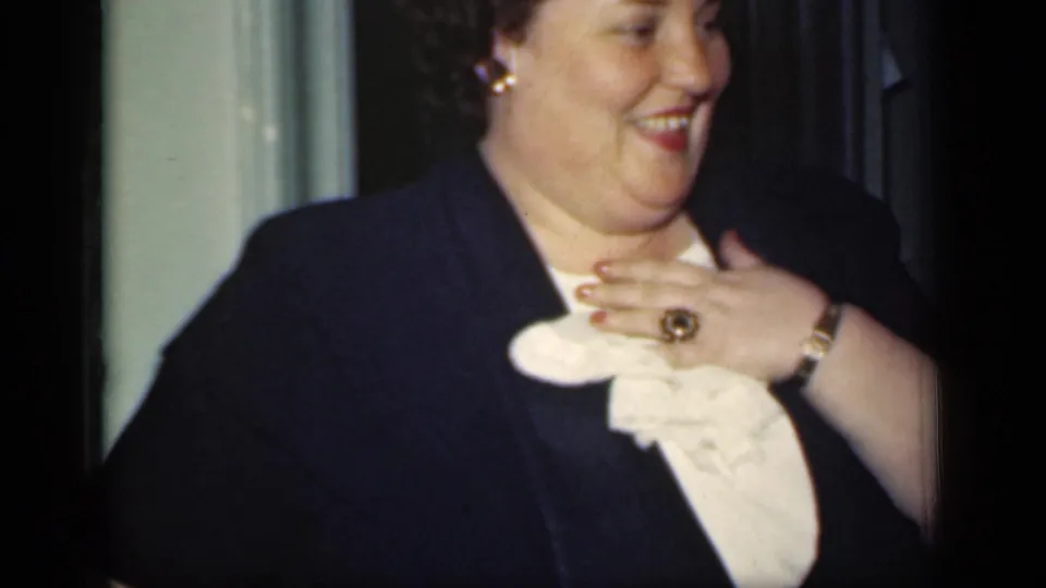A smiling woman in 1949 New York touches her white ruffled collar, with a gold ring, bracelet, dark jacket, and earrings visible.