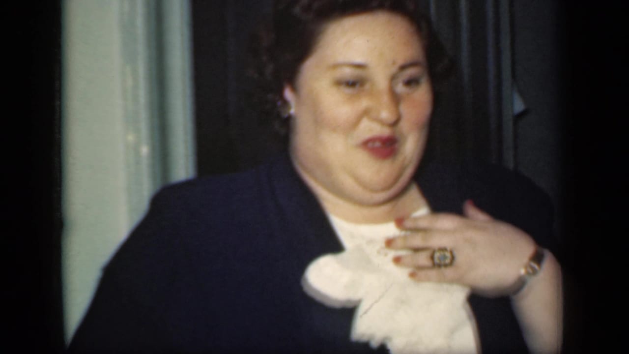 Woman in a dark suit smiles indoors with her hand near her chest.