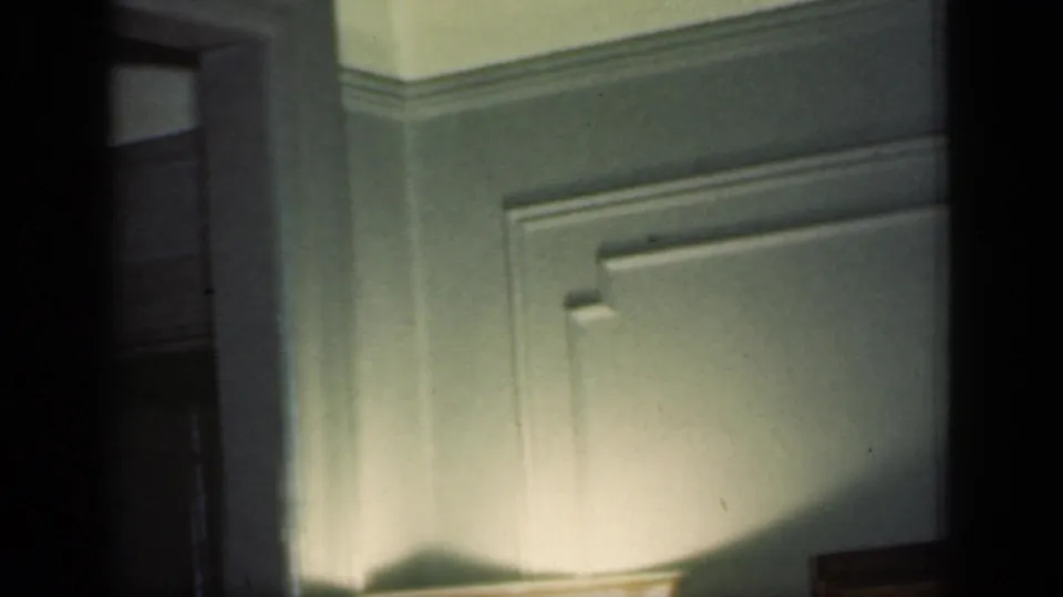 A 1949 home-movie frame lingers on a green interior corner with molding and a partially visible floor lamp.