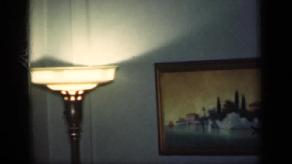 1949 New York 8mm frame showing a lit floor lamp casting warm light near a framed landscape painting in a private room.