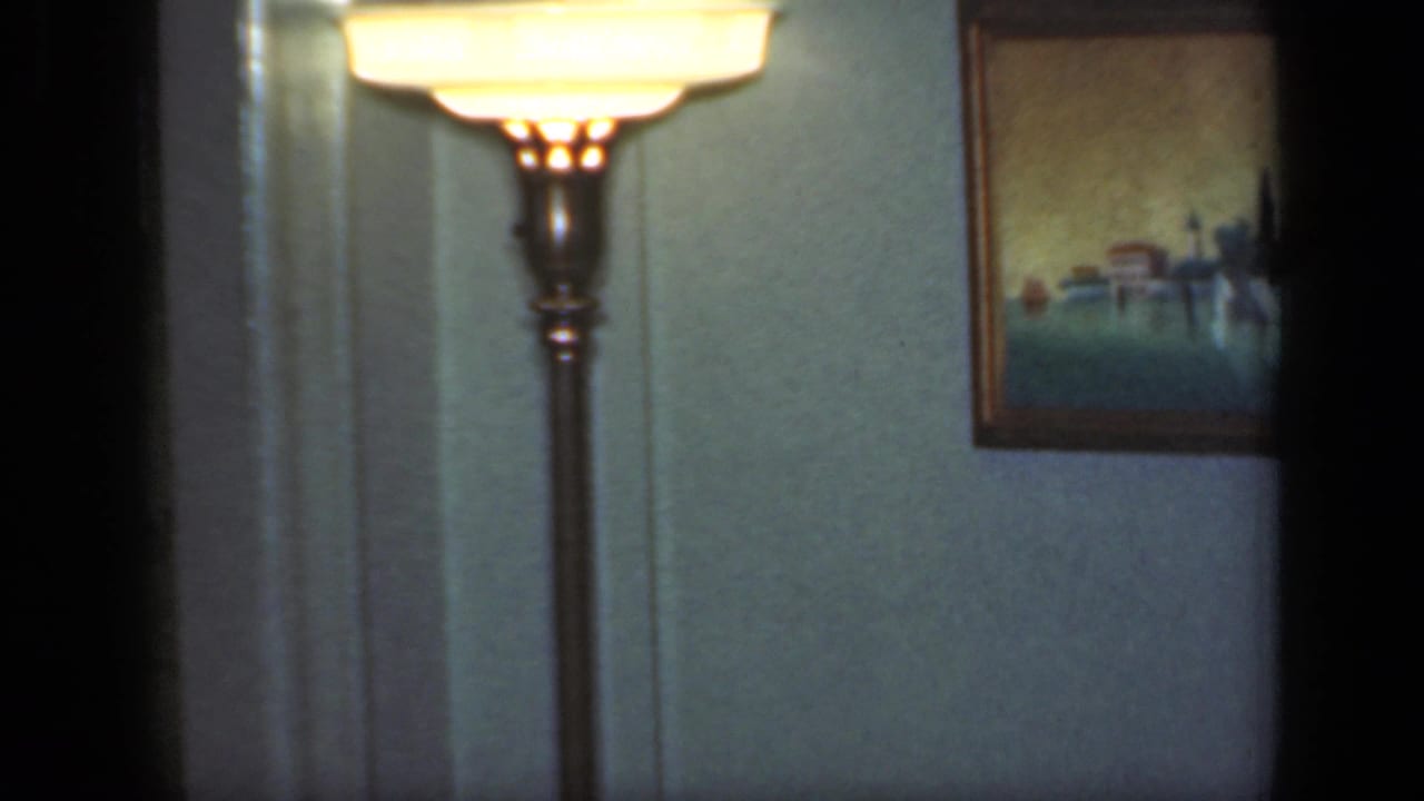 The camera holds on a lit floor lamp and framed wall picture in a 1949 New York private living-room interior.